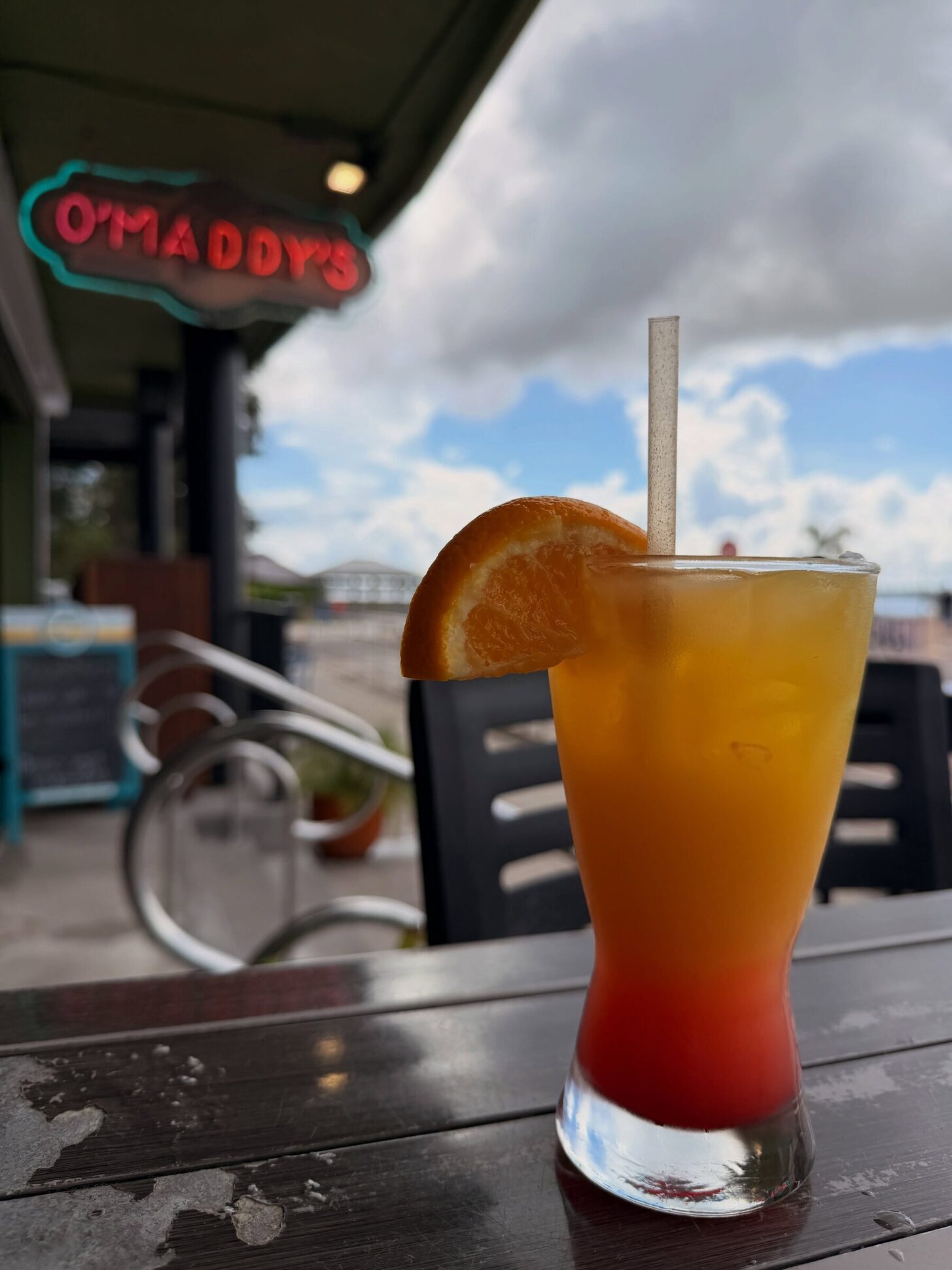 A tequila sunrise under the O'Maddy's neon sign, with the bay sky behind