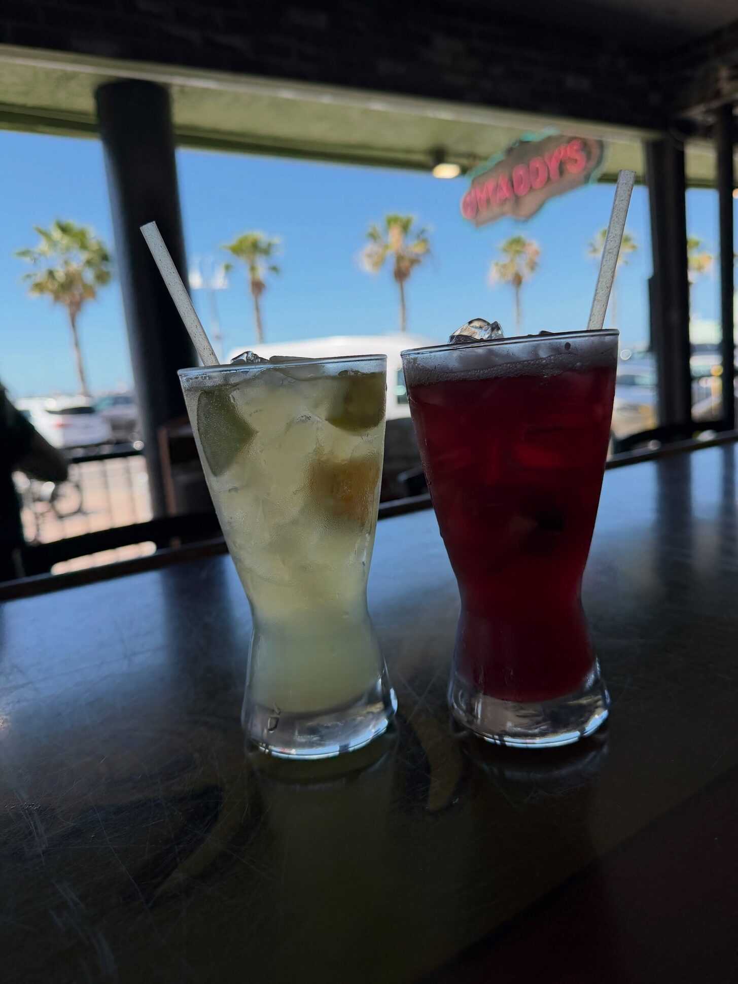Two cocktails on the O'Maddy's bar rail, with palms and the bay behind