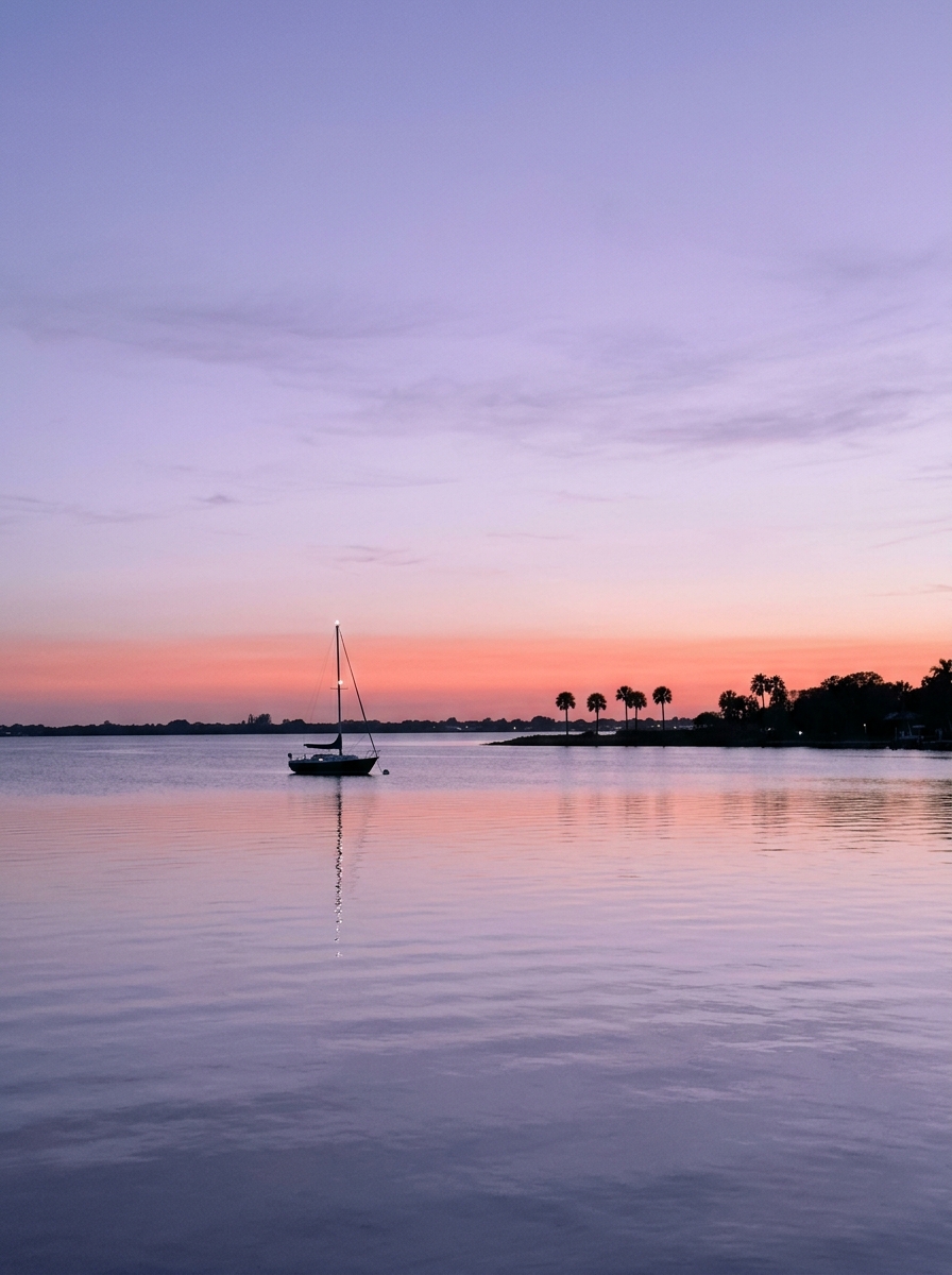 A lavender dusk sky over calm bay water with one anchored sailboat